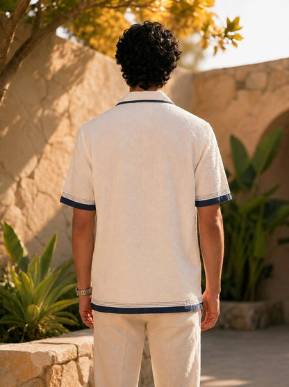 A man viewed from behind wearing a textured off-white short-sleeve polo shirt with navy blue trim on the collar, sleeve cuffs, and hem, paired with off-white trousers, set against a warm stone wall and green plants.