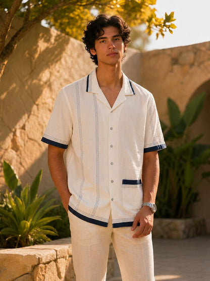 A man wearing a short-sleeve button-up camp shirt in off-white linen with navy blue trim, paired with matching off-white linen pants, standing outdoors in front of a stone wall and plants.